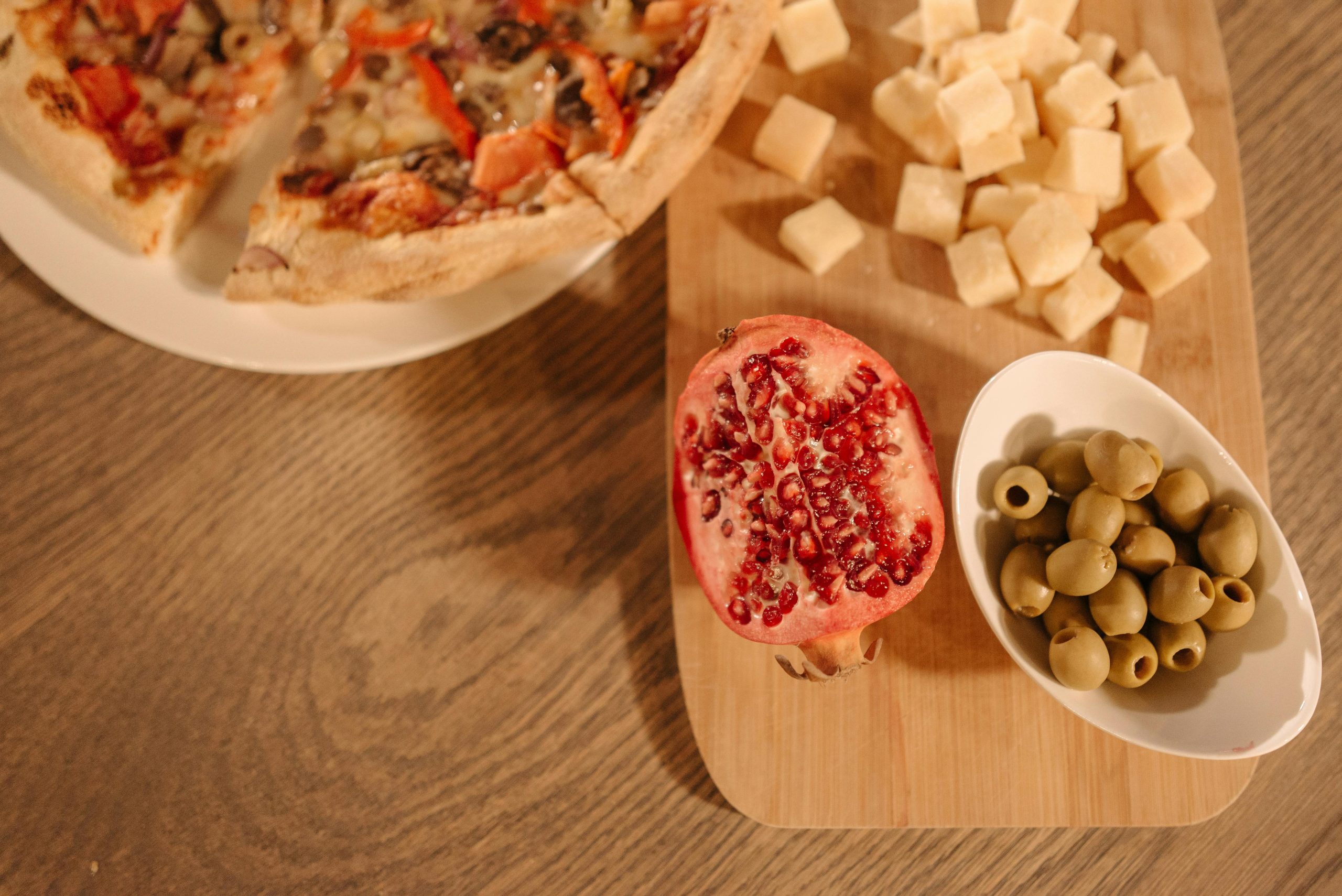A rustic spread featuring pizza, cheese cubes, pomegranate, and olives on a wooden board.